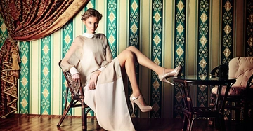 Frau in langem, beigem Kleid sitzt auf einem Stuhl mit einem Bein auf dem Tisch in einem Raum mit gemusterten Tapeten und Holzfußboden.