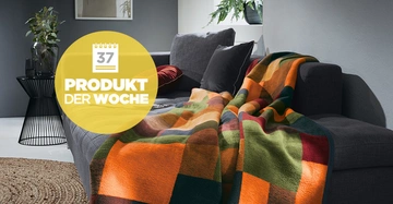 Eine Decke mit buntem Karomuster liegt auf einem grauen Sofa in einem modernen Wohnzimmer mit Pflanzen und Beistelltisch. Text: 37 PRODUKT DER WOCHE