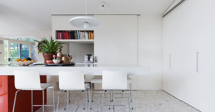 Weiße Stühle, die um einen weißen Tisch in einer modernen Küche mit weißer Wand und Regalen voller bunter Bücher stehen. Pflanzen und Haushaltsgeräte dekorieren den Tisch. Fenster im Hintergrund.