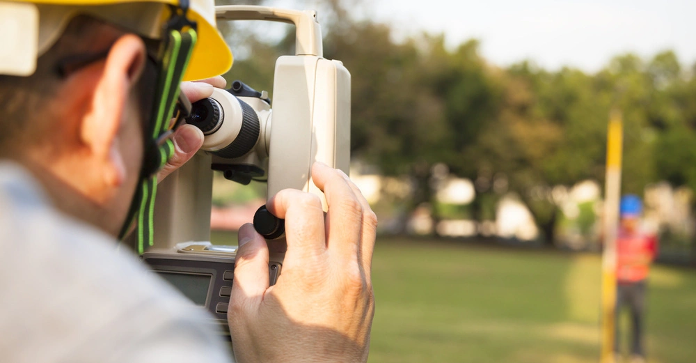 Ein Vermesser benutzt ein Theodolit zur Messung auf einem offenen Feld mit Bäumen im Hintergrund; im Hintergrund steht eine weitere Person mit einer Nivellierlatte.