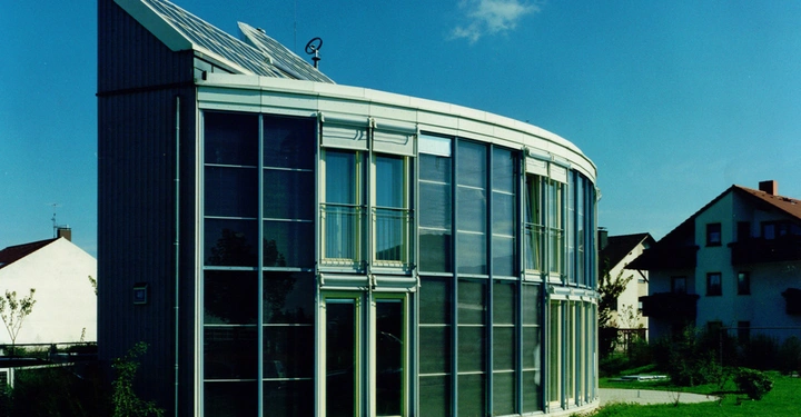 Ein modernes Glasgebäude steht auf einem grasbewachsenen Grundstück in einem Wohngebiet bei klarem sonnigen Himmel und angrenzenden traditionellen Häusern im Hintergrund.