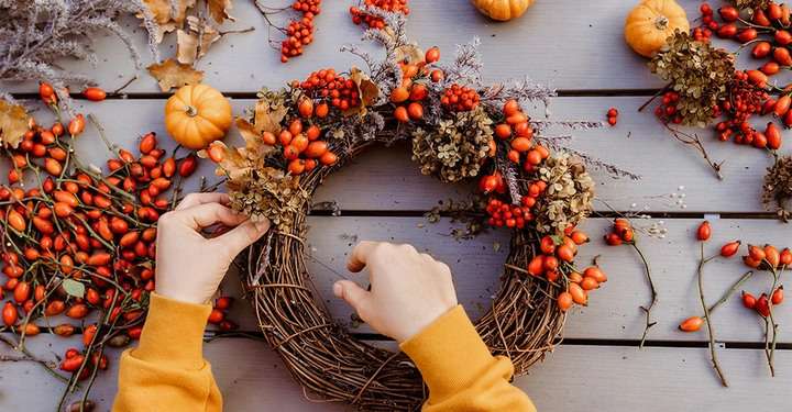 Frauenhände aus Vogelperspektive gestalten einen herbstlichen Türkranz mit Vogelbeeren, Zweigen und Minikürbissen. 
