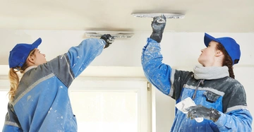 Zwei Personen mit blauen Kappen und Arbeitskleidung bearbeiten die Decke in einem Raum mir Spachtelwerkzeugen bei Tageslicht durch ein Fenster im Hintergrund.