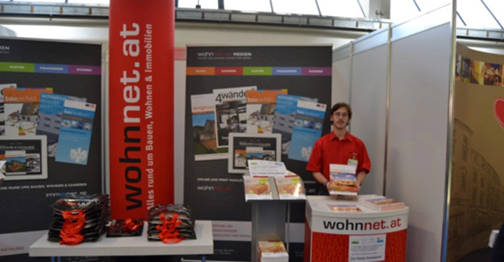 Galerie-Bild 3: Person steht an einem Informationsstand mit Stapeln von Broschüren vor großem roten Banner mit der Aufschrift wohnnet.at auf einer Messe oder Ausstellung umgeben von weiteren Informationsplakaten und Tischen mit Materialien.