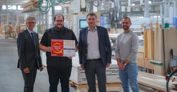 Vier Männer stehen in einer Fabrikhalle; der zweite von links hält ein Schild mit der Aufschrift MARKTTEST QUALITY AWARD 2022 TEST SIEGER HARTL HAUS 2022 Fertighaus MARKET INSTITUT