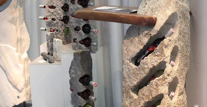 Galerie-Bild 8: Steinregale halten Weinflaschen in verschiedenen Aussparungen vertikal und horizontal angeordnet vor einem hellgrauen Vorhang umgeben von weiteren Weinflaschen und einem Holztisch.