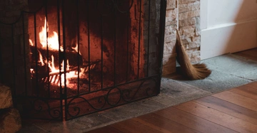 Loderndes Feuer brennt in einem Kamin hinter einem schwarzen Metallgitter. Eine Strohbesen steht neben dem gemauerten Kamin auf einem Holzboden in einer gemütlichen Raumumgebung.