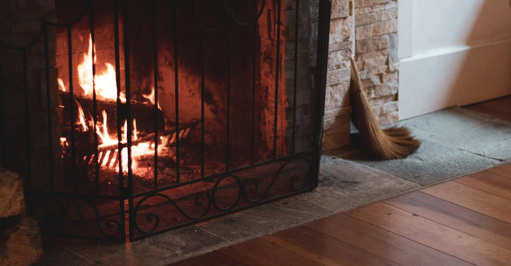 Loderndes Feuer brennt in einem Kamin hinter einem schwarzen Metallgitter. Eine Strohbesen steht neben dem gemauerten Kamin auf einem Holzboden in einer gemütlichen Raumumgebung.
