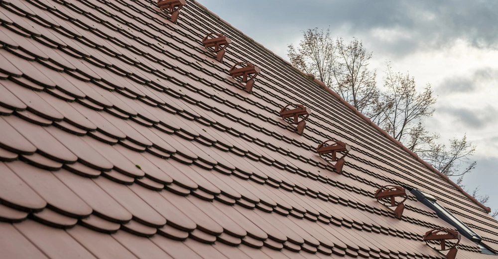 Dach mit Ziegeln bedeckt mehrere Schneebarrieren installiert um Schnee zu stoppen Baumzweige und bewölkter Himmel im Hintergrund.
