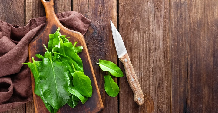 Frische grüne Blätter liegen auf einem Holzschneidebrett auf braunem Stoff eine Holzmesser liegt daneben alles auf einem rustikalen Holztisch.