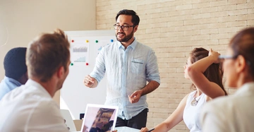 Ein Mann spricht zu einer Gruppe von vier Menschen sitzt an einem Tisch mit Laptops in einem modernen Büro mit Flipchart im Hintergrund.