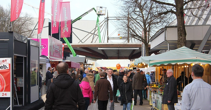 Galerie-Bild 1: Messebesucher gehen durch eine belebte Messehalle mit Ständen und bunten Fahnen im Hintergrund einige Bäume Blätterlose Bäume flankieren den Weg Werbebanner Text http www.sez-sonnenschutz.at
