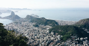 Panoramablick auf eine Stadt mit vielen Hochhäusern verteilt zwischen grünen Hügeln und umgeben von Küste und Meer im Hintergrund einige Inseln und Berge am Horizont