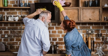 Ein Mann und eine Frau reinigen zusammen eine Küche mit Backsteinwand die Frau wischt ein Regal ab während der Mann die Dunstabzugshaube abwischt zahlreiche Küchenutensilien stehen im Hintergrund.