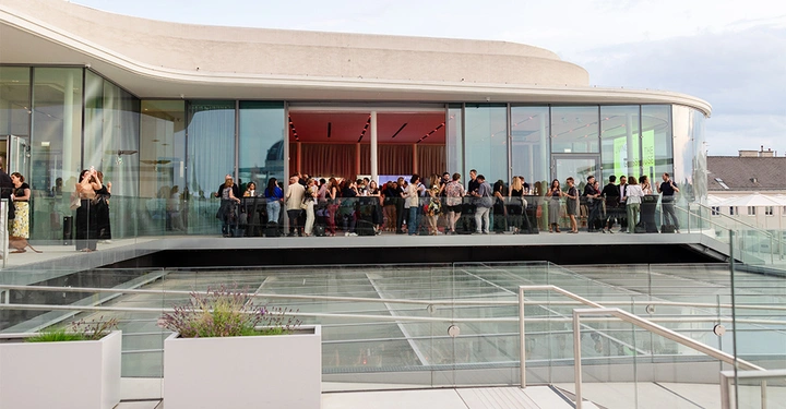 Frontalansicht der Libelle, Restaurant auf dem Dach des Leopold Museums in Wien. Glasdach und Glasfassade, Treppenabgang im Vordergrund, Menschen tummeln sich auf der Terrasse