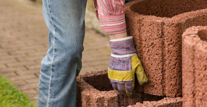 Eine Person mit gestreiftem Hemd und Jeans hebt orangefarbene Betonsteine im Freien an trägt Handschuhe und steht auf einem gepflasterten Weg neben einer Grasfläche