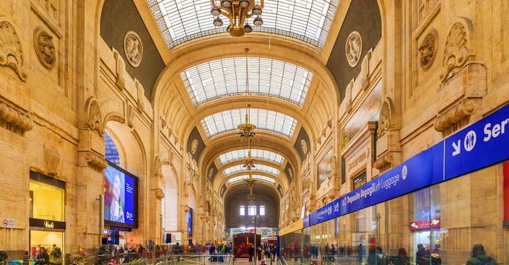 Menschen gehen durch eine große historische Bahnhofshalle mit hohen Decken und dekorativen Bögen. Riesenbildschirme und geschäftige Geschäfte umgeben die Fläche. Blaues Schild sagt Deposito Bagagli/Left Luggage mit Pfeil.