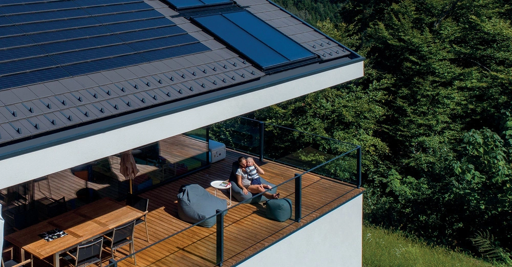 Eine Person sitzt mit einem Kind auf einem Holzdeck im Freien neben einem kleinen runden Tisch und Sitzsäcken. Das darüberliegende Dach ist mit Solarpanels bedeckt. Der Hintergrund zeigt grüne Bäume.