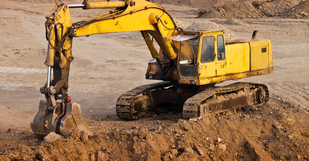 großer gelber Bagger auf leerem Areal beim Aushub einer Baugrube, dominierende Farben sind gelb und braun. 