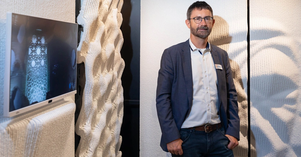 Mann steht in einem Raum zwischen strukturierten weißen Wänden und einem Flachbildfernseher der ein architektonisches Design zeigt. Der Mann trägt eine Brille ein Hemd und ein Sakko.