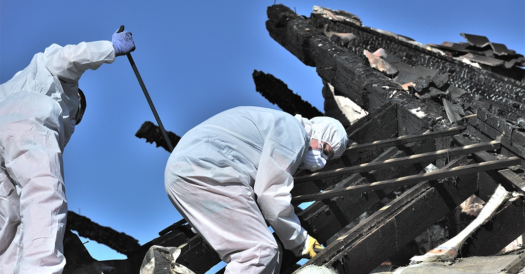 Verkohlter Dachstuhl, zwei vermummte Brandsanierer arbeiten in weißen Schutzanzügen und mit Maske