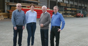 Vier Personen stehen nebeneinander auf einem gepflasterten Hof vor einem Industriegebäude mit gestapeltem Holz. Im Hintergrund sind Gabelstapler zu sehen. Sie tragen legere Kleidung.