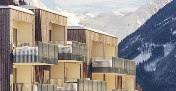 Mehrstöckige Wohngebäude mit Balkonen, auf denen Schnee liegt, stehen vor einer Kulisse aus verschneiten Bergen unter klarem Himmel.