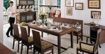 Frau trägt einen Teller in einem elegant gedeckten Esszimmer mit Holzmöbeln. Der Raum ist hell, mit großen Fenstern und blühenden Blumen auf dem Tisch.