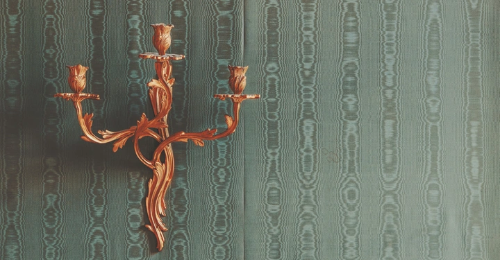 Ein goldener, verzierter Kerzenhalter mit drei Armen ist an einer Wand mit grün-grau gemustertem Tapetenmuster befestigt