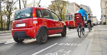 Ein rotes Auto fährt auf einer Stadtstraße zwei Radfahrer sind nebeneinander unterwegs im Hintergrund sind Bäume Gebäude und ein Straßenbahn zu sehen auf der Straße steht ENDE.