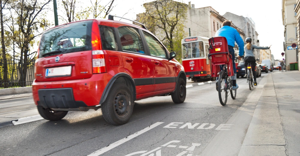 Ein rotes Auto fährt auf einer Stadtstraße zwei Radfahrer sind nebeneinander unterwegs im Hintergrund sind Bäume Gebäude und ein Straßenbahn zu sehen auf der Straße steht ENDE.