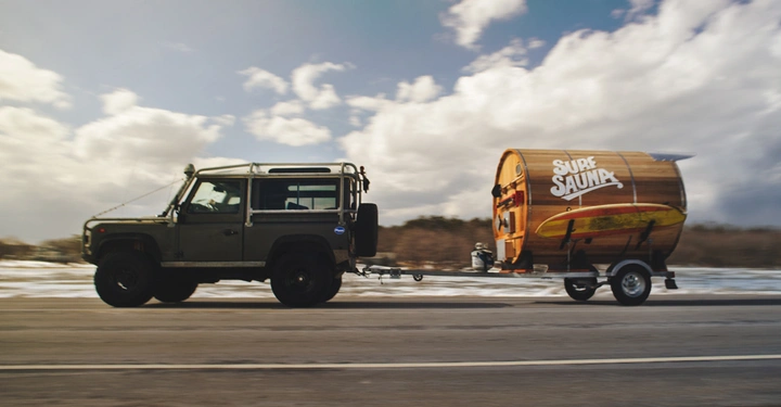 Ein grüner Geländewagen zieht einen hölzernen Saunawagen mit der Aufschrift Surf Sauna auf einer Landstraße bei bewölktem Himmel.