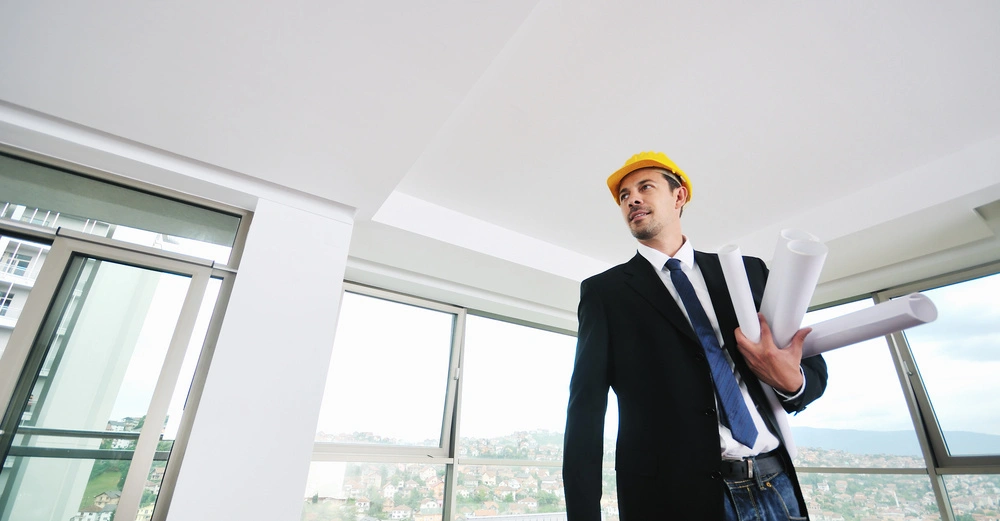 Ein Mann trägt einen gelben Bauhelm und hält mehrere Baupläne. Er steht in einem modern ausgestatteten Raum mit großen Fenstern, durch die man eine städtische Landschaft sieht.