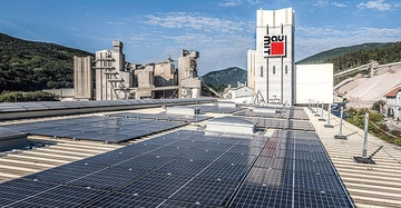 Solarmodule bedecken das Dach eines Industriegebäudes im Vordergrund während im Hintergrund ein Fabrikkomplex mit hohen Silos und Schornsteinen vor bewaldeten Hügeln steht. Das Logo "Baumit" ist zu sehen.