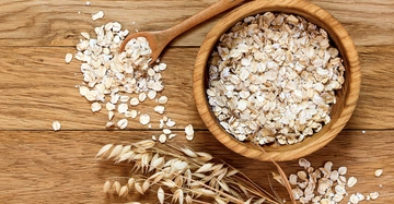 Eine Holzschale voller Haferflocken eine Handvoll Haferflocken auf einem Holzlöffel und einige Haferähren liegen auf einem Holztisch.