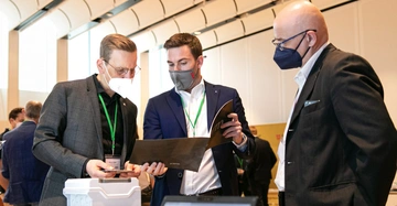 Drei maskierte Männer betrachten in einem modernen Konferenzraum gemeinsam ein Dokument. Zwei stehen, einer hält das Dokument, und weitere Personen sind im Hintergrund erkennbar. Ein Erkennungsanhänger enthält den Text Moritz Hüther.