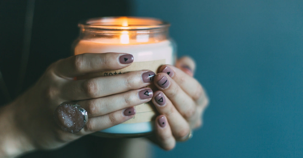 Hände halten eine brennende Kerze in einem Glas in der Nahaufnahme Nagellack mit Mustern Ring sichtbar Hintergrund dunkel und unscharf