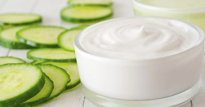 Galerie-Bild 6: Eine Schüssel mit weißer Creme steht auf einem Holztisch umgeben von frischen Gurkenscheiben. Im Hintergrund ist ein weiteres Gefäß mit einer grünen Flüssigkeit zu sehen.