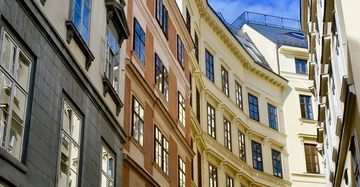 Mehrstöckige Gebäude stehen nebeneinander in einer engen europäischen Straße Die Fenster reflektieren den blauen Himmel und die gegenüberliegenden Fassaden Hellgelbe und orangefarbene Fassaden vor einem klaren Himmel