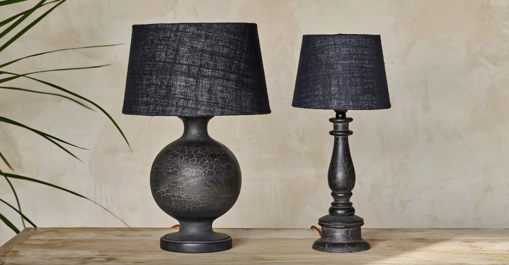 Zwei schwarze Tischlampen stehen auf einem Holztisch links steht eine breite Lampe mit rundem Fuß und rechts eine schmalere Lampe mit gedrehtem Fuß beide vor beigem Hintergrund.