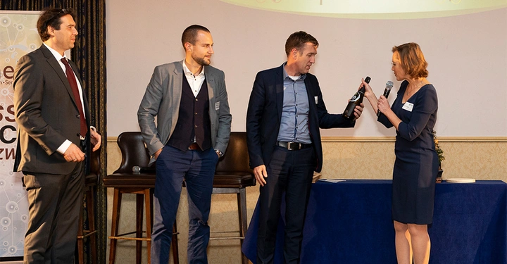 Galerie-Bild 4: Vier Menschen in formeller Kleidung auf einer Bühne Ein Mann rechts überreicht einer Frau eine Weinflasche Zwei weitere Männer stehen links in der Nähe eines Banners mit der Aufschrift Edge Services und BAAS STÜTZEN.