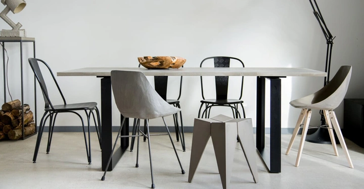 Ein Esstisch mit einer Holzschale darauf, umgeben von verschiedenen modernen Stühlen, steht in einem minimalistischen, hellen Raum mit einem Kaminholzstapel und einer hohen Lampe.