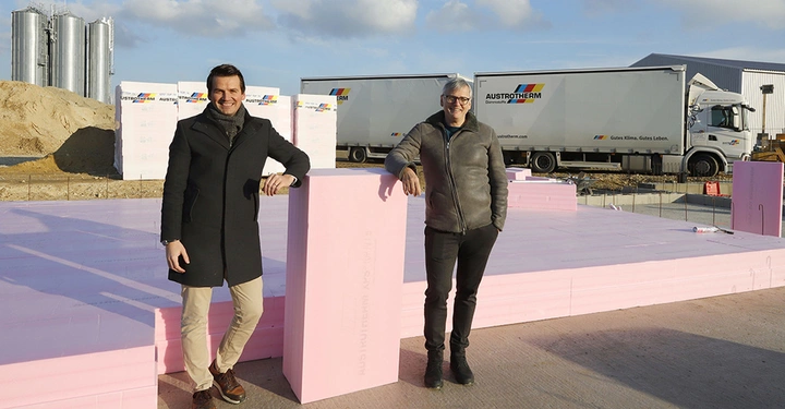 Zwei Männer stehen auf einer Baustelle mit rosa Isolierplatten. Im Hintergrund befinden sich Sandhaufen, LKWs mit der Aufschrift "Austrotherm" und ein Gebäude.