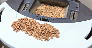 Große Menge an Pellets liegt auf einer weißen Oberfläche vor einem schwarzen Behälter mit einem offenen Fach voller Pellets in einem hellen Raum