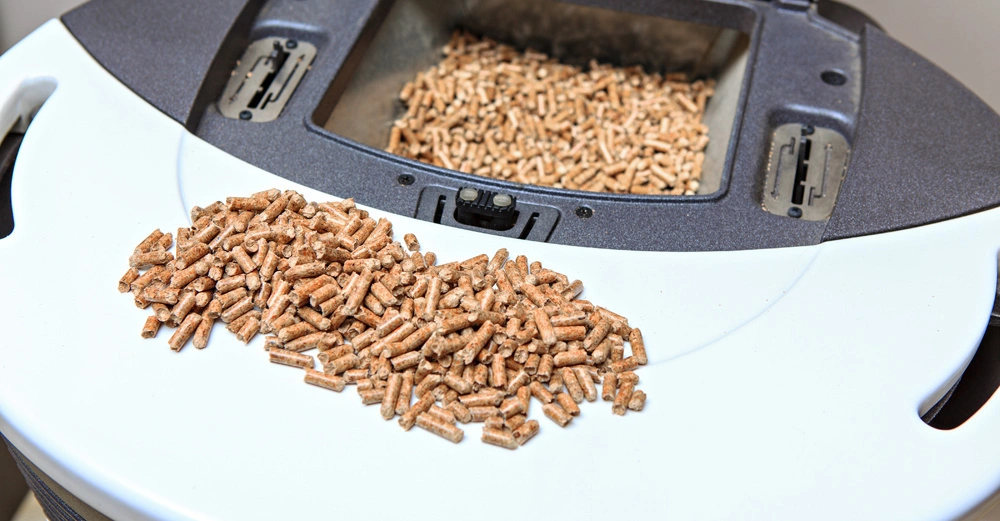 Große Menge an Pellets liegt auf einer weißen Oberfläche vor einem schwarzen Behälter mit einem offenen Fach voller Pellets in einem hellen Raum