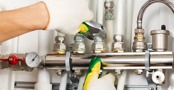 Hände mit Arbeitshandschuhen benutzen einen Schraubenschlüssel um Rohrverbindungen zu fixieren in einem technischen Wartungsbereich mit mehreren Rohren und Ventilen. Ein Manometer ist sichtbar.