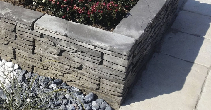 Galerie-Bild 9: Steinmauer umgibt eine Fläche mit blühenden roten und weißen Pflanzen innerhalb eines gepflasterten Außenbereichs zwei Arten von Steinen liegen am Fuß der Mauer weiß und grau