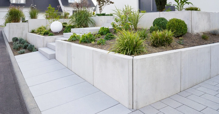 Ein Garten mit mehreren Betonstufen ist bepflanzt mit verschiedenen Sträuchern und Gräsern. Im Hintergrund ist ein modernes Haus mit großen Fenstern im Wohnviertel zu sehen.