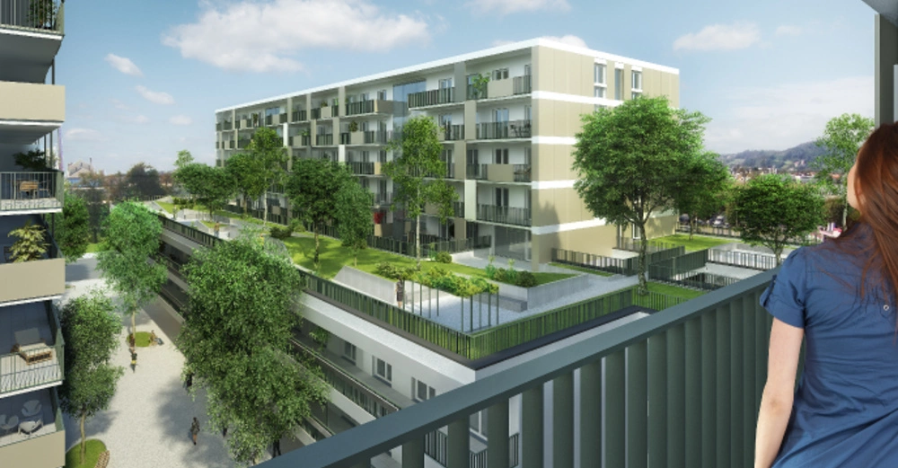 Eine Frau steht auf einem Balkon und blickt auf einen modernen Wohnkomplex mit mehreren Stockwerken und Balkonen umgeben von vielen Bäumen und einem grünen Innenhof bei Tageslicht.