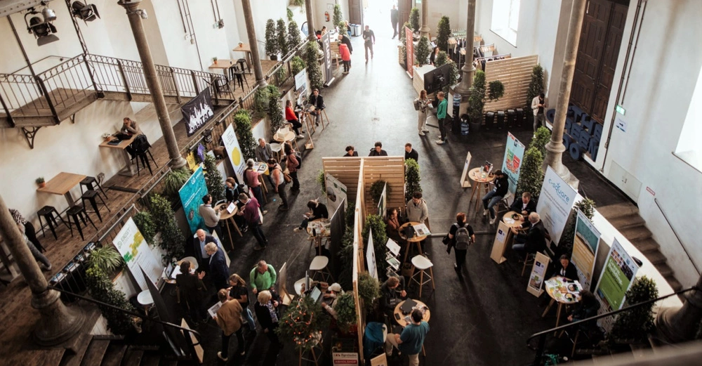 Menschen interagieren an verschiedenen Ständen; es gibt Plakate und Tische innerhalb einer großen Halle mit hohen Decken und Säulen einige Menschen sitzen an erhöhten Tischen auf der oberen Etage.
